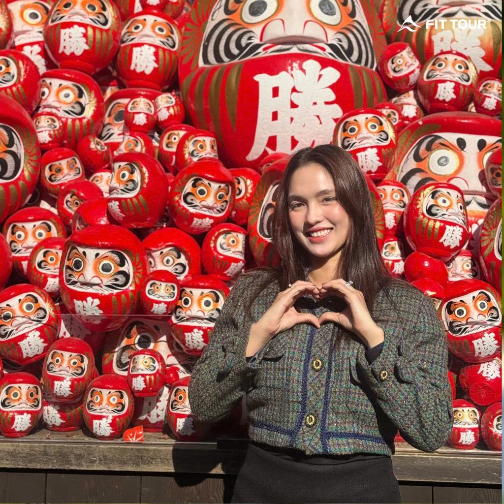 Khách hàng nữ rạng rỡ tạo dáng trái tim trước rừng búp bê Daruma đỏ rực, một điểm check-in “hot” trong tour.